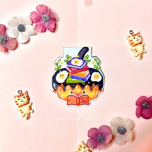 May include: A colorful bookmark featuring a cartoon crab holding a book titled "Guide to Being a Bookworm". The crab is sitting in a frying pan with a stack of books and two fried eggs.