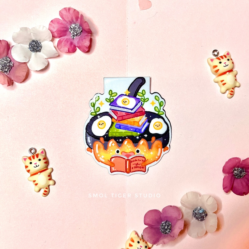 May include: A colorful bookmark featuring a cartoon crab holding a book titled "Guide to Being a Bookworm". The crab is sitting in a frying pan with a stack of books and two fried eggs.