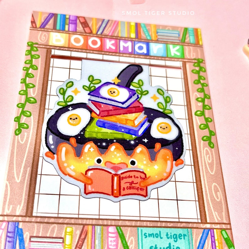 May include: A colorful bookmark featuring a cartoon crab holding a book titled "Guide to be a Calicifer". The crab is sitting in a frying pan with a stack of books and eggs on top. The bookmark is surrounded by a wooden frame with a bookshelf design.