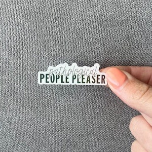 May include: A rectangular sticker with the text "pathological PEOPLE PLEASER" in black and white. The word "pathological" is in a cursive font, and the rest of the text is in a sans-serif font. The sticker is held by a hand with painted nails.