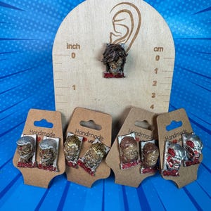 May include: Handmade earrings featuring a detailed face design with red accents. The earrings are displayed on small brown cards with the word "Handmade" printed on them. A wooden ear-shaped display with inch and cm measurements is also shown.