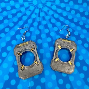 May include: A pair of rectangular drop earrings featuring a metallic gray finish with a central circular cutout. Each earring has four gold-colored accents positioned around the inner circle and a textured top edge, suspended from silver-toned hooks.