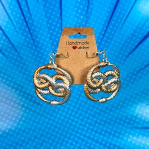May include: A pair of silver and gold snake earrings. The earrings are in the shape of two snakes intertwined, forming a circle. The earrings are hanging from a brown card with the text "Handmade with love".