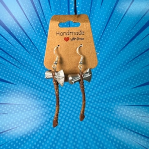 May include: A pair of silver earrings shaped like Thor's hammer, Mjolnir, with brown leather straps hanging from the bottom. The earrings are displayed on a brown card with the text "Handmade with love".