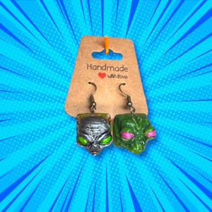 May include: A pair of handmade alien head earrings. One earring is silver with green eyes, the other is green with pink accents. The earrings hang from silver hooks and are displayed on a brown tag with the text "Handmade with love".