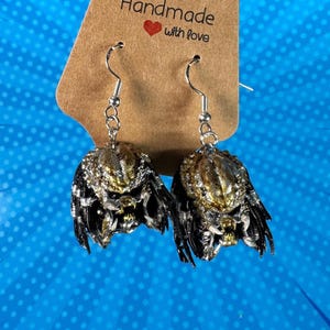 Puede incluir: Un par de pendientes colgantes plateados con un diseño de Depredador. Los pendientes tienen un acabado metálico detallado con detalles dorados, plateados y negros. Cuelgan frente a una etiqueta marrón con las palabras "Handmade with love" y un corazón rojo.