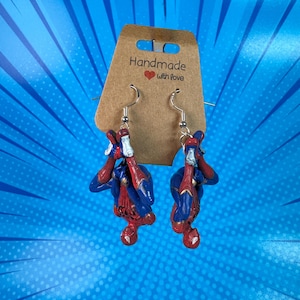 Puede incluir: Un par de pendientes de plata con una figura de Spiderman roja, azul y dorada colgando boca abajo. Los pendientes están en una tarjeta marrón con el texto "Handmade with love".