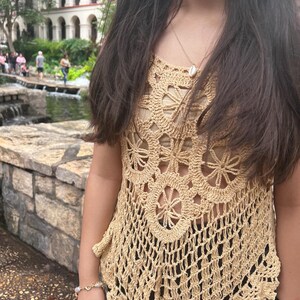 May include: A woman wearing a beige crochet top with a shell necklace. The top has a scalloped hem and a delicate floral pattern.