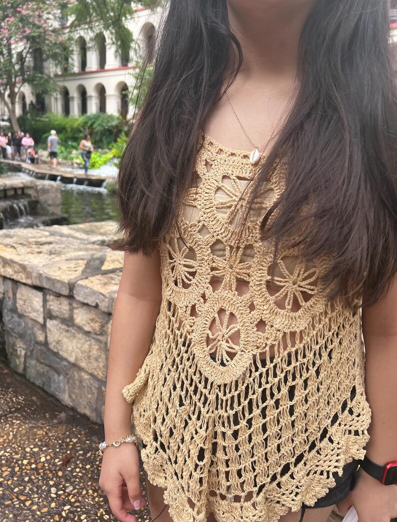 May include: A woman wearing a beige crochet top with a shell necklace. The top has a scalloped hem and a delicate floral pattern.