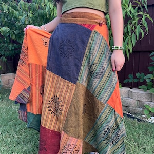 May include: A long, multi-colored patchwork wrap skirt with a brown suede waistband. The skirt features a variety of printed designs, including suns, moons, and swirls. The skirt is worn with a green crop top.