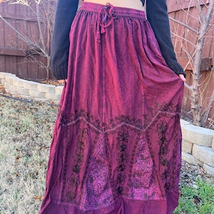 May include: A long, flowing burgundy skirt with an elasticated waistband and drawstring. The skirt features intricate black embroidered floral patterns along the hemline. The fabric has a textured appearance. The skirt is worn outdoors.