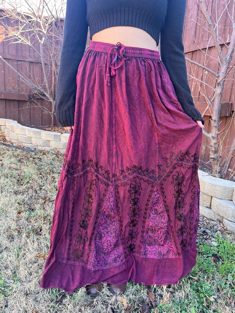 May include: A long, flowing burgundy skirt with an elasticated waistband and drawstring. The skirt features intricate black embroidered floral patterns along the hemline. The fabric has a textured appearance. The skirt is worn outdoors.