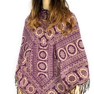 K&ouml;nnte beinhalten: Lila und cremefarbener Poncho mit Fransenbesatz. Der Poncho hat ein geometrisches Muster mit kreisf&ouml;rmigen Motiven.