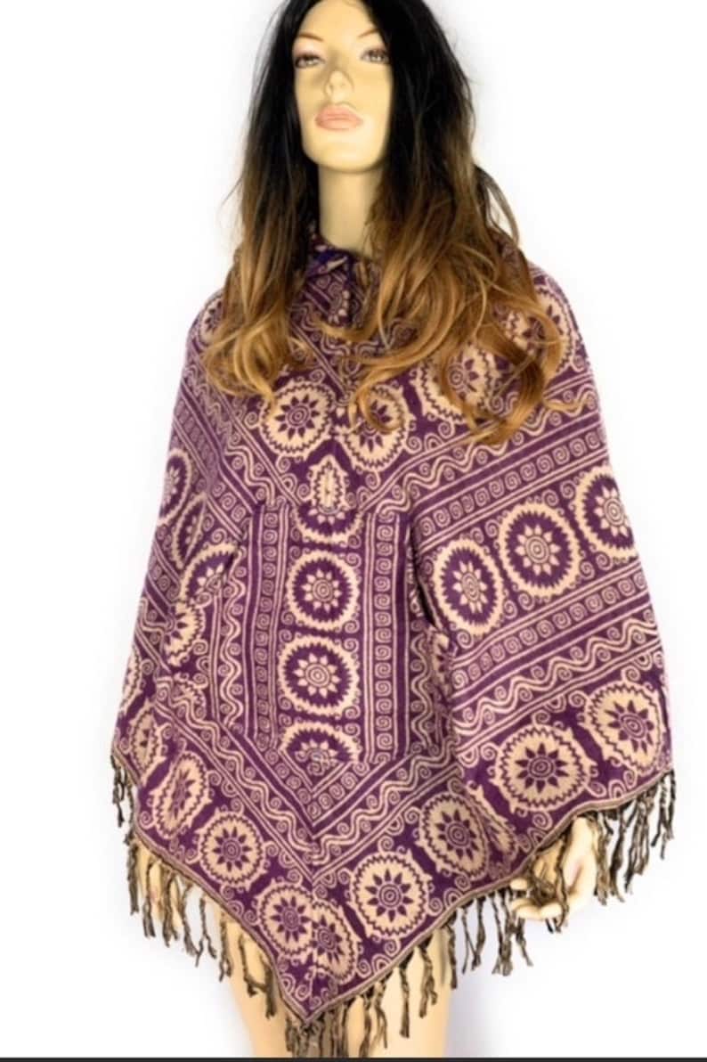 K&ouml;nnte beinhalten: Lila und cremefarbener Poncho mit Fransenbesatz. Der Poncho hat ein geometrisches Muster mit kreisf&ouml;rmigen Motiven.