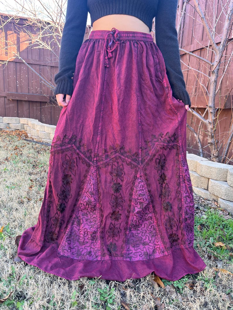 May include: A long, flowing burgundy skirt with detailed embroidered floral patterns. The skirt has a drawstring waist and a tiered hemline. The fabric looks to be a soft, textured material, creating a bohemian aesthetic.