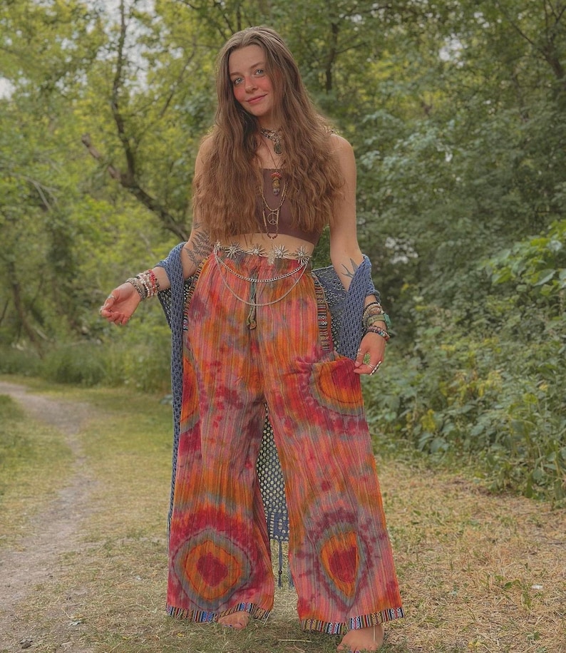 Puede incluir: Una mujer con un top marr&oacute;n corto, un cintur&oacute;n plateado y pantalones tie-dye. Est&aacute; de pie en un entorno de bosque y tiene el pelo largo y casta&ntilde;o.