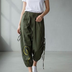 May include: Olive green cropped pants featuring an elastic waistband with a drawstring, side pockets, and adjustable side ties. The pants display a yellow spiral graphic design on the lower right leg and are paired with a plain white t-shirt.
