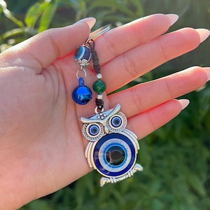 Könnte beinhalten: Ein silberner Eulen-Schlüsselanhänger mit blauem und weißem Augendesign. Der Schlüsselanhänger hat einen blauen Glockenanhänger und grüne Perlen. Der Eulenanhänger ist mit blauen und weißen Akzenten versehen.