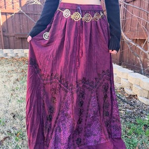 May include: A long, flowing burgundy skirt with detailed embroidered patterns. The skirt has a drawstring waist and is worn with a gold-coloured belt with spiral designs. The outfit is completed with a black long-sleeved top.