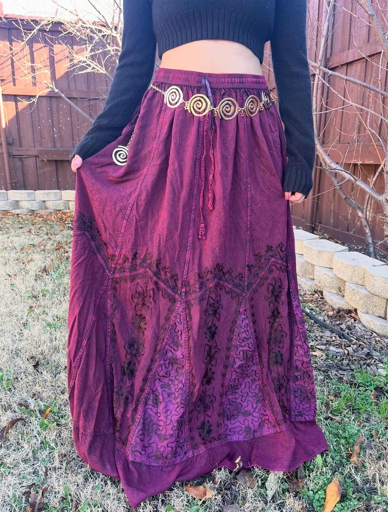 May include: A long, flowing burgundy skirt with detailed embroidered patterns. The skirt has a drawstring waist and is worn with a gold-coloured belt with spiral designs. The outfit is completed with a black long-sleeved top.
