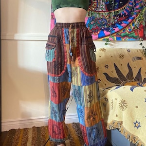 Puede incluir: Pantalones de patchwork multicolores con cintura elástica y dos bolsillos grandes. El diseño presenta paneles marrones, azules, naranjas y rojos con patrones de sol y abstractos. Los pantalones son de tela suave.