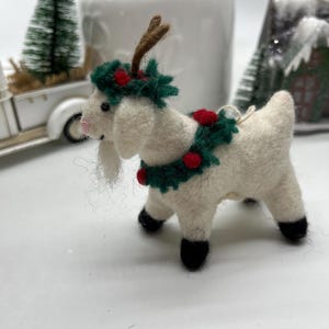 May include: A white felt goat Christmas ornament with a green and red wreath around its neck and brown antlers. The goat has black hooves and a pink nose. A small white string is attached for hanging.
