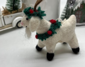 May include: A white felt goat Christmas ornament with a green and red wreath around its neck and brown antlers. The goat has black hooves and a pink nose. A small white string is attached for hanging.