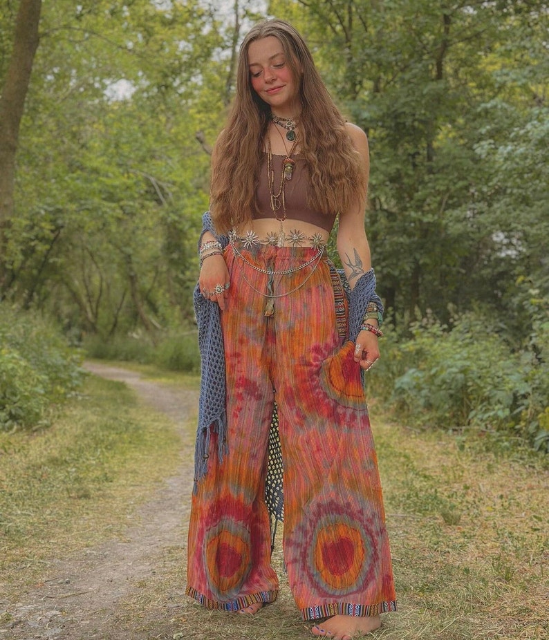 Puede incluir: Una mujer con un top marr&oacute;n corto, un cintur&oacute;n de cadena plateada con abalorios y pantalones tie-dye. Est&aacute; de pie en un entorno de bosque.