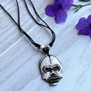 May include: A white skull pendant necklace with a black cord. The skull is carved from wood and has a detailed design. The necklace is accented with black and white beads.