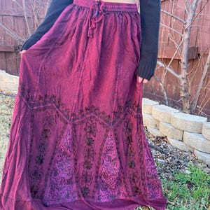 May include: A long, flowing burgundy skirt with an elasticated waistband and drawstring. The skirt features intricate dark floral embroidery along the hemline and panels. The fabric has a slightly textured appearance.