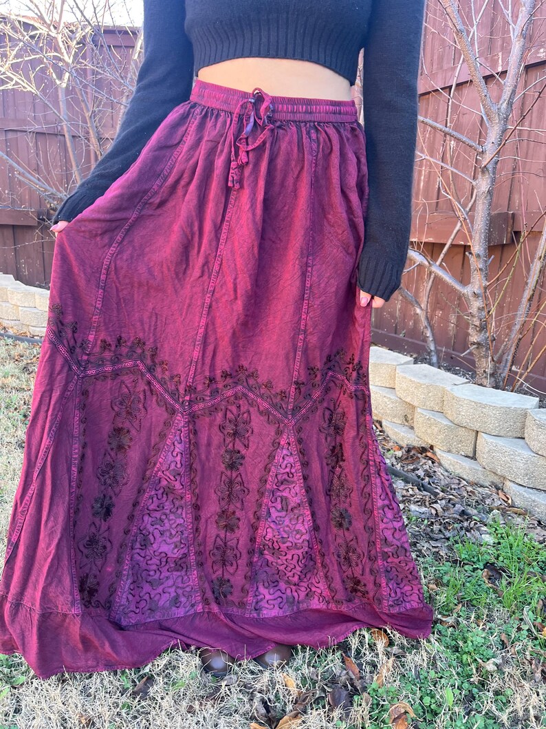 May include: A long, flowing burgundy skirt with an elasticated waistband and drawstring. The skirt features intricate dark floral embroidery along the hemline and panels. The fabric has a slightly textured appearance.