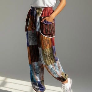 May include: Loose-fitting trousers featuring a patchwork design with various rectangular panels in shades of red, blue, brown, and green. The pants include a drawstring waistband, two large front pockets with white trim, and black sun and floral motifs.
