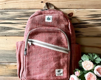 May include: A pink hemp backpack with a zipped pocket and a small green leaf logo. The backpack is sitting on a wooden surface with a bouquet of pink flowers.