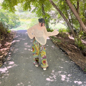 Puede incluir: Una persona con un su&eacute;ter de crochet blanco y pantalones tie-dye camina por un camino pavimentado en un &aacute;rea boscosa.