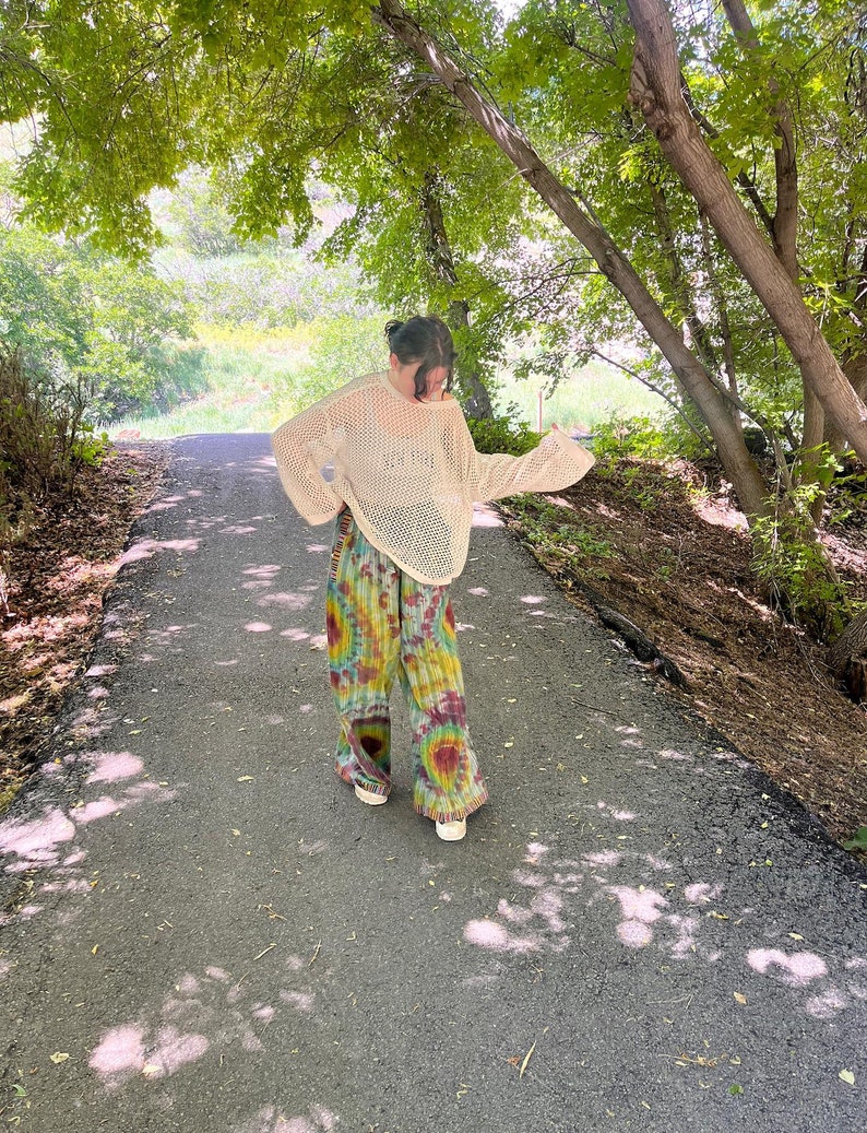 Puede incluir: Una persona con un su&eacute;ter de crochet blanco y pantalones tie-dye camina por un camino pavimentado en un &aacute;rea boscosa.