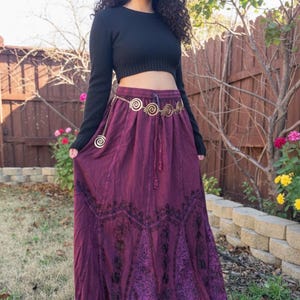May include: A woman in a black crop top and a long, flowing, deep purple skirt with black embroidered patterns. A gold-coloured chain belt cinches the waist. The background features a wooden fence and greenery. The skirt's length is approximately 100 cm.