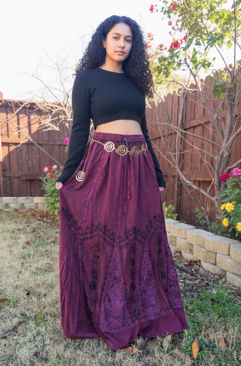May include: A woman in a black crop top and a long, flowing, deep purple skirt with black embroidered patterns. A gold-coloured chain belt cinches the waist. The background features a wooden fence and greenery. The skirt's length is approximately 100 cm.