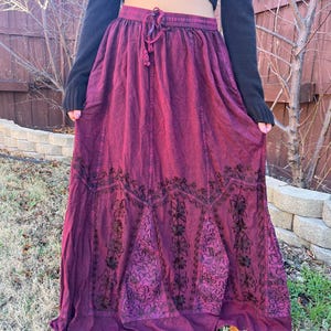 May include: A long, flowing burgundy skirt with an embroidered floral design. The skirt has a drawstring waist and a slightly faded appearance. The skirt is made of a soft, flowing fabric, perfect for a bohemian or casual style.