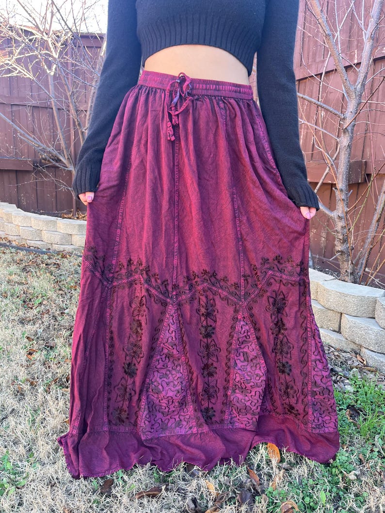 May include: A long, flowing burgundy skirt with an embroidered floral design. The skirt has a drawstring waist and a slightly faded appearance. The skirt is made of a soft, flowing fabric, perfect for a bohemian or casual style.