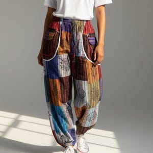 Peut inclure: Pantalon ample en patchwork composé d'un mélange vibrant de panneaux de tissu rouge, bleu, orange et marron. Le pantalon comprend une taille élastique, deux poches latérales avec bordure blanche et des motifs de soleil et lune imprimés en noir.