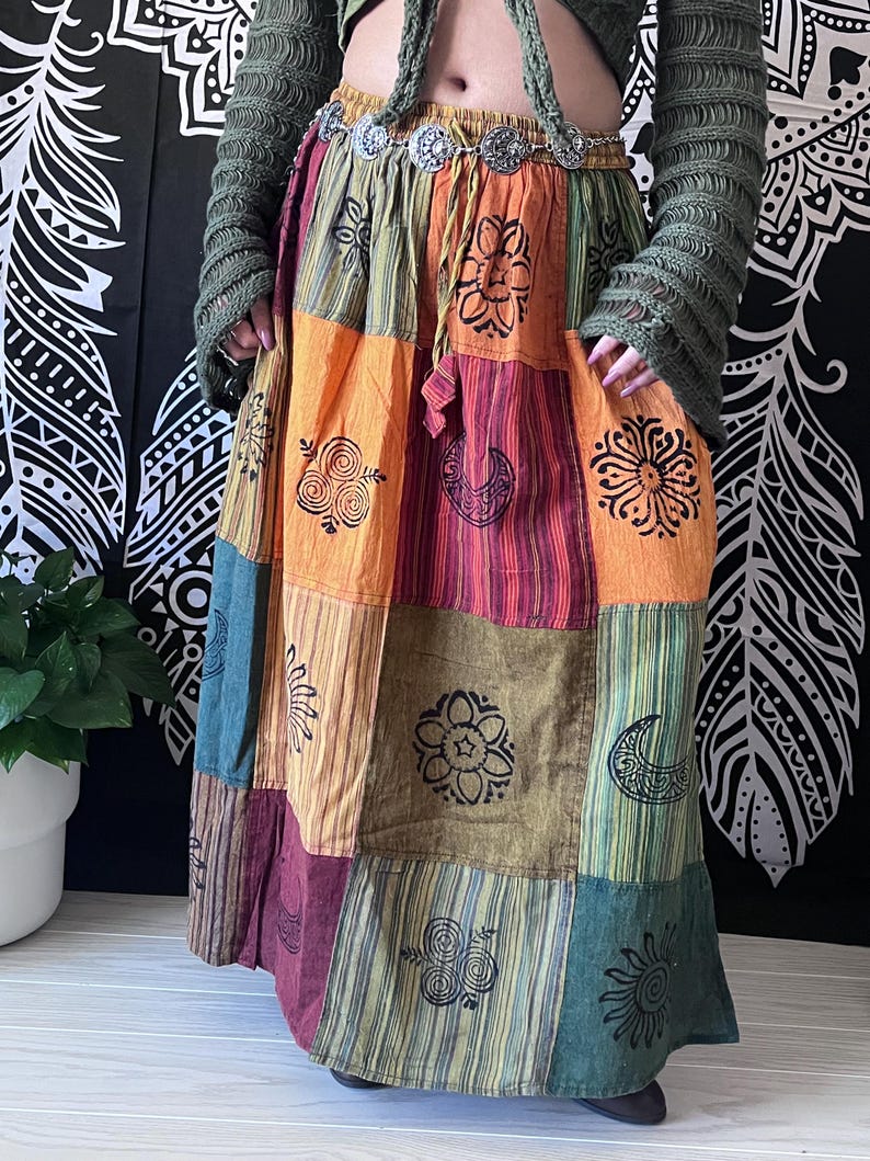 Puede incluir: Una falda larga de retazos multicolores con una variedad de dise&ntilde;os impresos, incluyendo flores, lunas y espirales. La falda tiene una cintura con un cintur&oacute;n plateado.