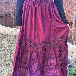 May include: A long, flowing burgundy skirt with an embroidered floral design. The skirt has a drawstring waist and a darker, decorative pattern near the hem. The fabric appears to be a soft, textured material.