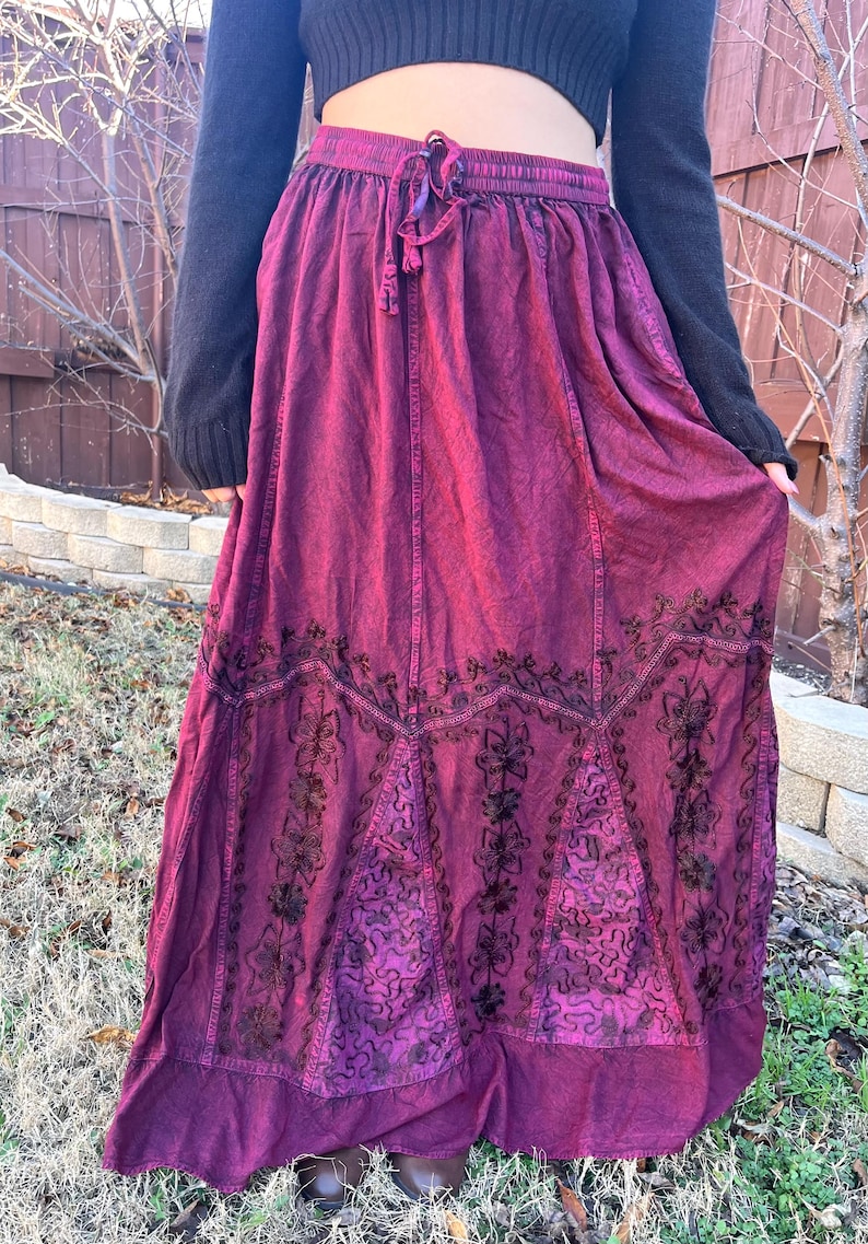 May include: A long, flowing burgundy skirt with an embroidered floral design. The skirt has a drawstring waist and a darker, decorative pattern near the hem. The fabric appears to be a soft, textured material.