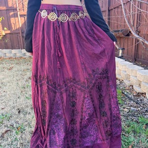 May include: A long, flowing burgundy skirt with embroidered floral patterns. The skirt has a drawstring waist and is paired with a gold-coloured belt with spiral designs. The outfit is completed with a black long-sleeved top.