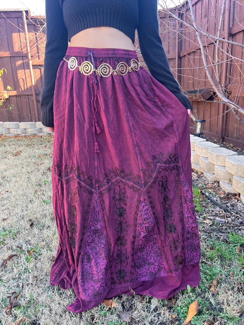 May include: A long, flowing burgundy skirt with embroidered floral patterns. The skirt has a drawstring waist and is paired with a gold-coloured belt with spiral designs. The outfit is completed with a black long-sleeved top.