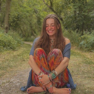 Puede incluir: Una joven con el pelo largo y casta&ntilde;o se sienta con las piernas cruzadas en un camino en un bosque. Lleva una blusa de ganchillo azul y blanca, pantalones coloridos y varias pulseras. Sonr&iacute;e y tiene los ojos cerrados.