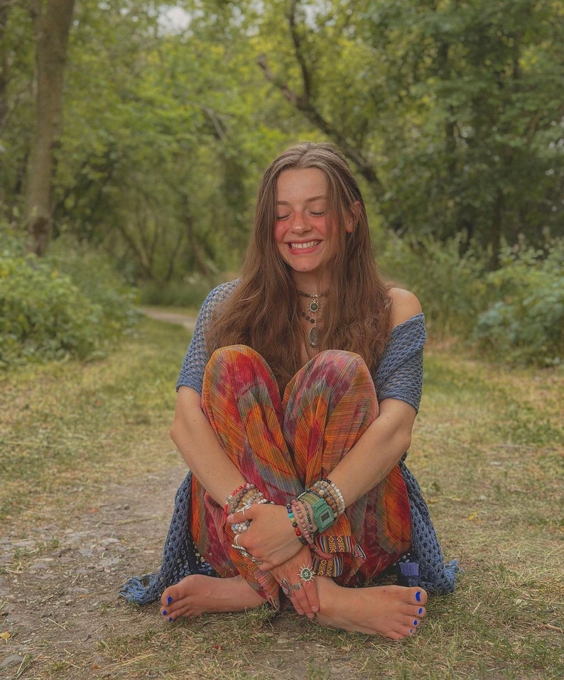 Puede incluir: Una joven con el pelo largo y casta&ntilde;o se sienta con las piernas cruzadas en un camino en un bosque. Lleva una blusa de ganchillo azul y blanca, pantalones coloridos y varias pulseras. Sonr&iacute;e y tiene los ojos cerrados.
