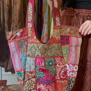 以下が含まれることがあります： 赤いベースにカラフルな刺繍が施されたパッチワークのトートバッグ。バッグは大きく開いたトップと2本の長いストラップが付いています。