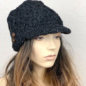 May include: A dark gray, knitted newsboy cap with a small brim. The cap features a textured pattern and is adorned with two brown buttons on the side. The hat is designed to be worn on the head.