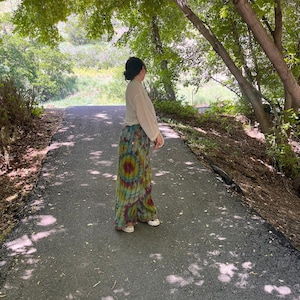 Puede incluir: Una persona con un top de crochet blanco y pantalones tie-dye se encuentra en un camino pavimentado en un &aacute;rea boscosa. El camino est&aacute; bordeado de &aacute;rboles y vegetaci&oacute;n verde.